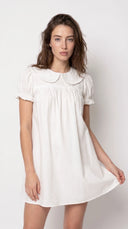 A woman modeling a short-sleeve white babydoll dress with a ruffled Peter Pan collar and gathered smocked bodice, photographed against a plain off-white studio background.