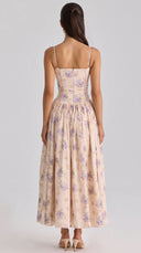 A woman viewed from the back wearing a sleeveless maxi dress with thin straps, a fitted bodice, and a full gathered skirt in a light blush pink floral print with purple flowers, paired with beige high heels against a light gray studio background.