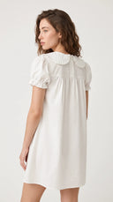 A woman modeling a short-sleeve white dress with a ruffled Peter Pan collar, shown from a three-quarters back view against a plain light gray studio background.