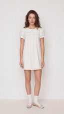 A woman modeling a short-sleeved white dress with a Peter Pan collar, standing against a plain off-white studio background.